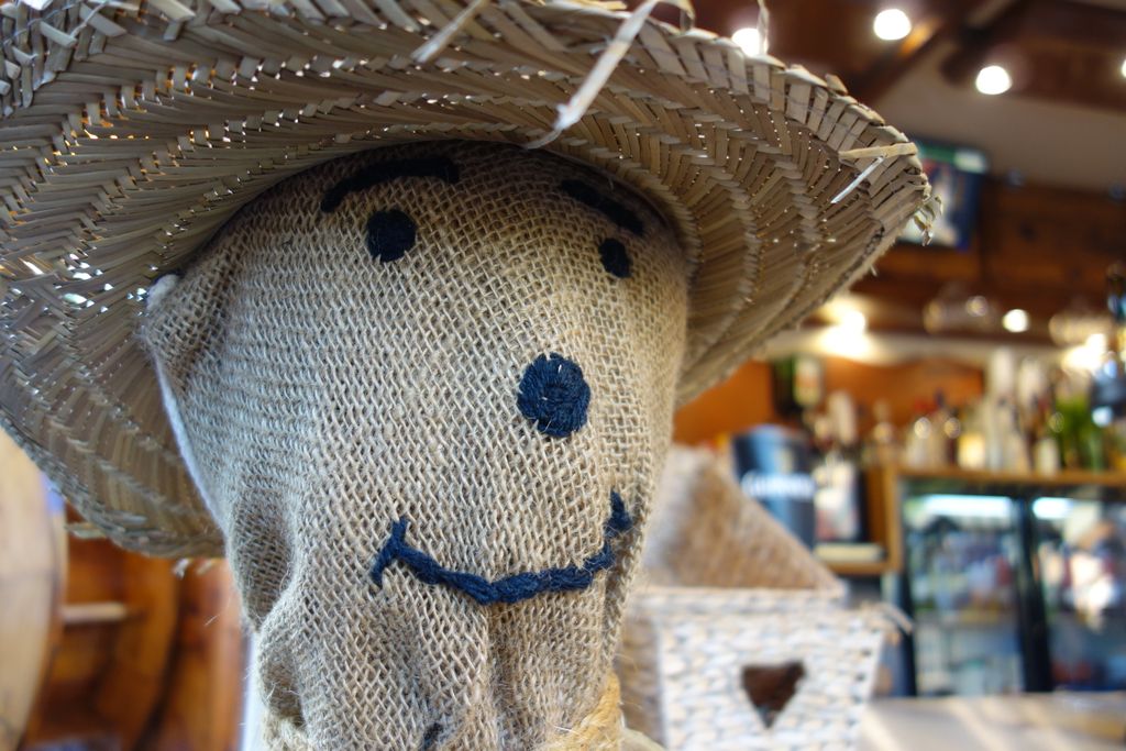 Smiling burlap scarecrow with straw hat, rustic indoor setting.